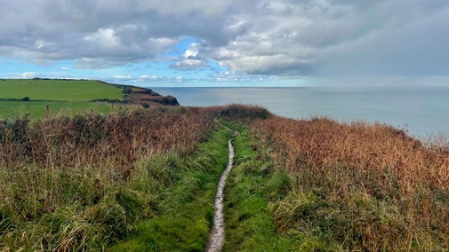 A coastal cliff top path with cloudy skies and views of a calm sea during autumn.